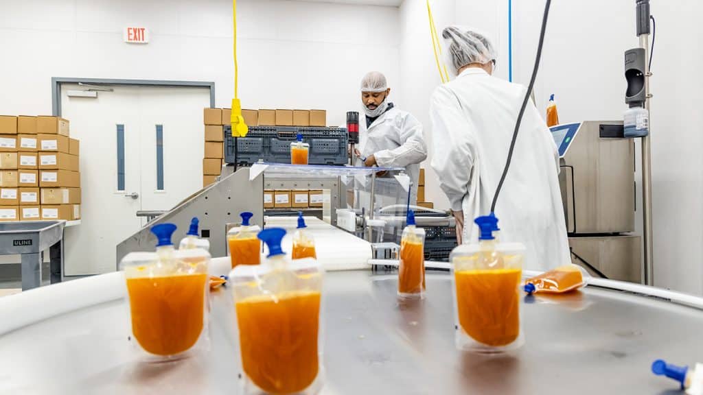 Two people in white lab coats and hair nets work at machinery. Packets of orange puree are in the foreground.