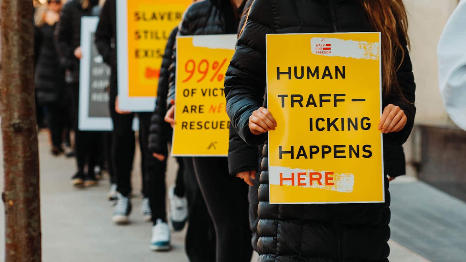 a line of protesters in dark clothing carry yellow signs that read "human trafficking happens here"