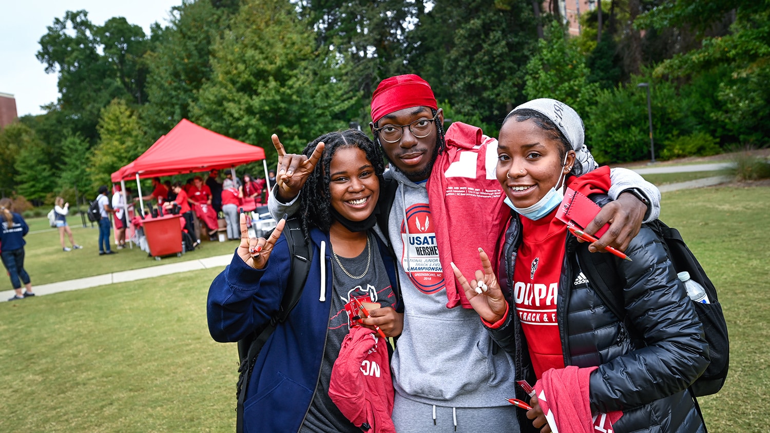 Three students pose for a photo holding NC State swag that they got from the Red and White Week kickoff event.