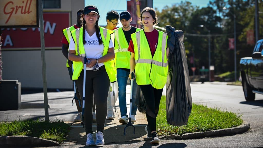 A group of five students wearing safety vests walk down Hillsborough Street collecting garbage.