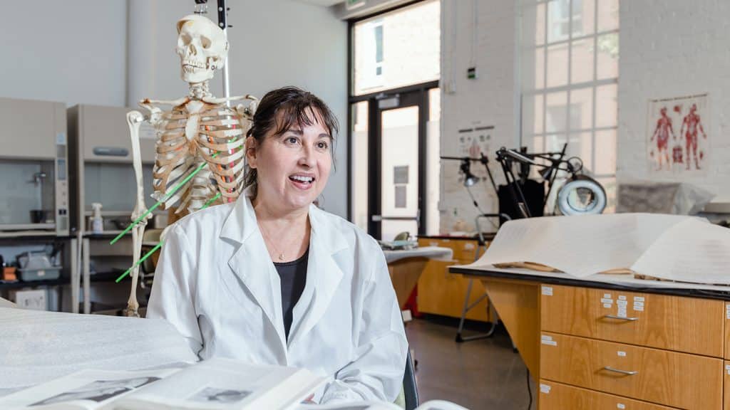 Ann Ross sits at a table in her lab. Textbooks are open on the table, and an upright skeleton stands behind her. 