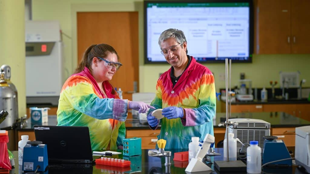 Carlos Goller, an associate teaching professor in the Biotechnology Program, and Madison Routh, a post-baccalaureate researcher, wear rainbow-colored lab coats as they work together in a campus lab space.