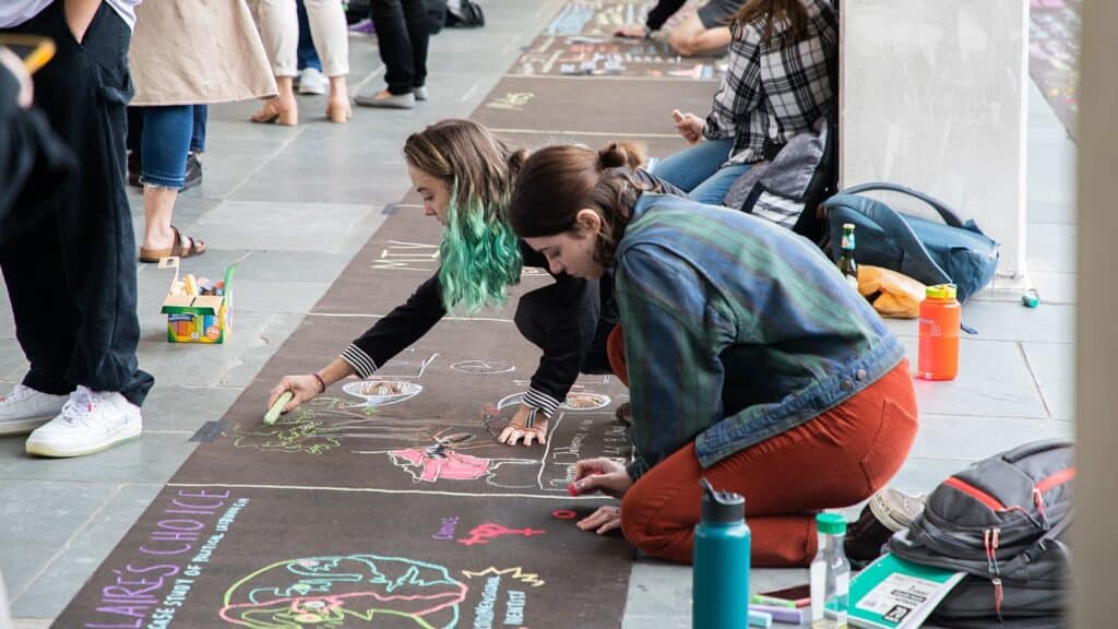 Two students draw art with sidewalk chalk outside of D.H. Hill Library; the illustrations are related to their research projects.