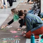 Two students draw art with sidewalk chalk outside of D.H. Hill Library; the illustrations are related to their research projects.