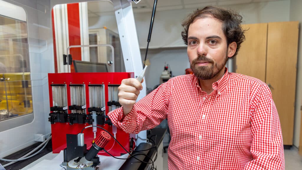 Nathan Woodward stands next to a small red machine in Amassian's lab. Woodward holds a microscope slide with tweezers. 