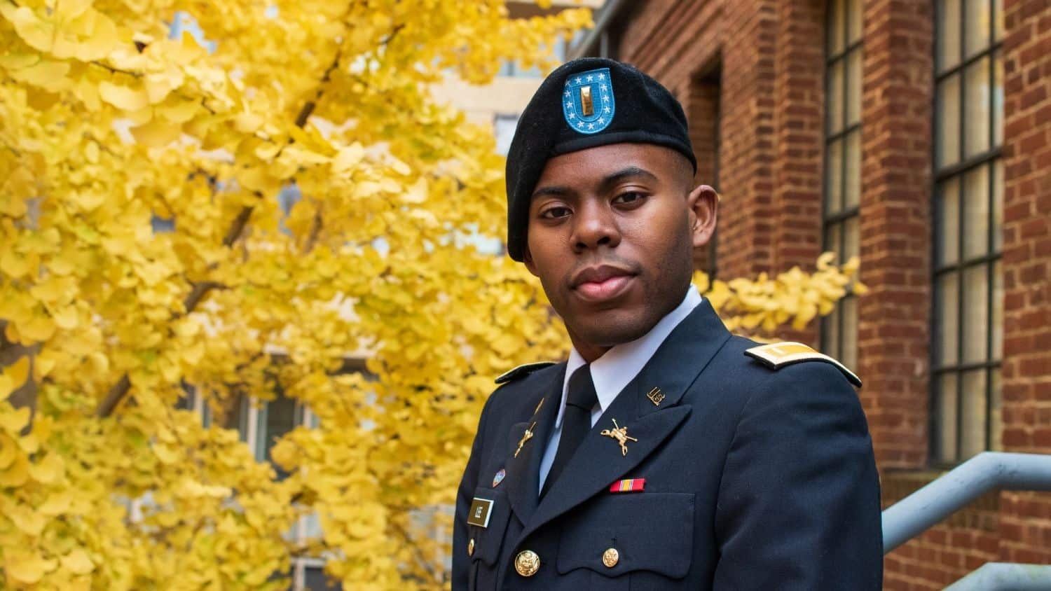 Young alumnus poses in a military uniform in front of a brick building and foliage.