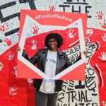A student holding a frame that reads "#FirstInThePack" in front of a mural.
