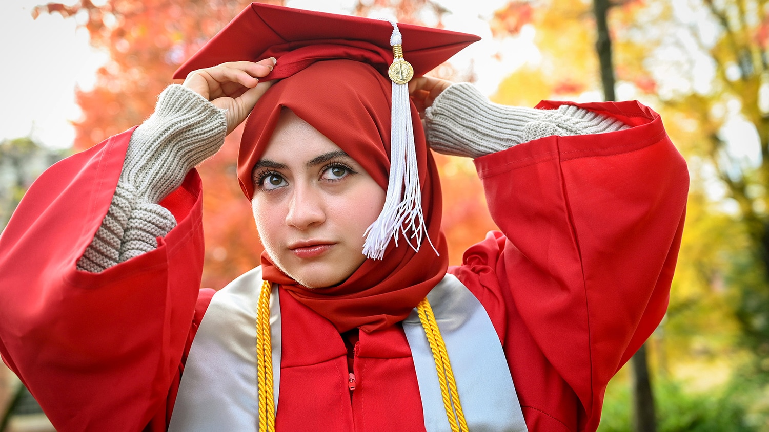 Portrait of Maab Aldulimy wearing her red graduation gown and adjusting her cap and tassel.
