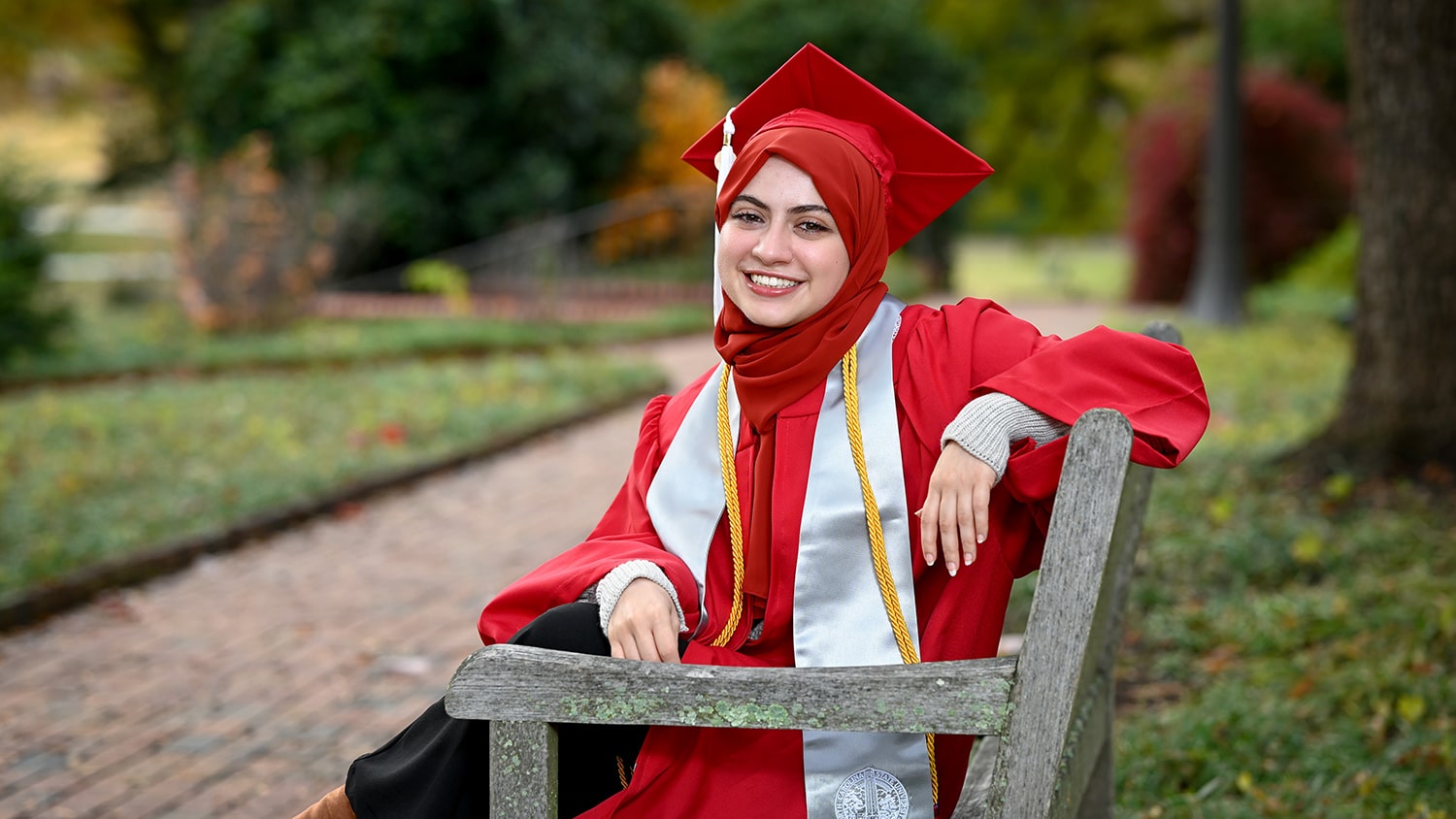 Maab Aldulimy sits on a bench while wearing her red cap and gown.