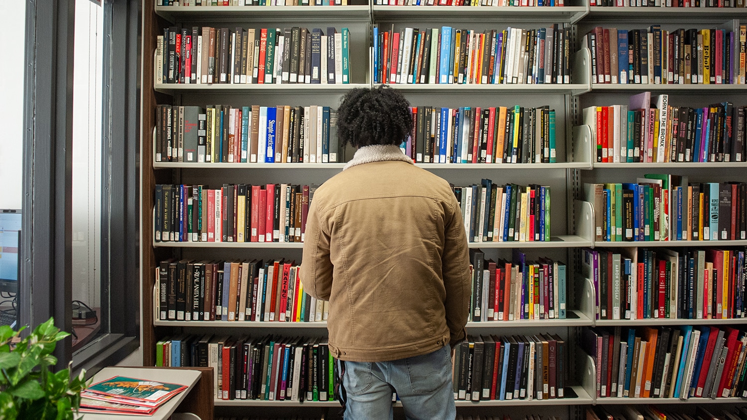 A student stands in front of a multi-tiered library book shelf. Hundreds of books, all of different sizes and colors, are on the shelf.