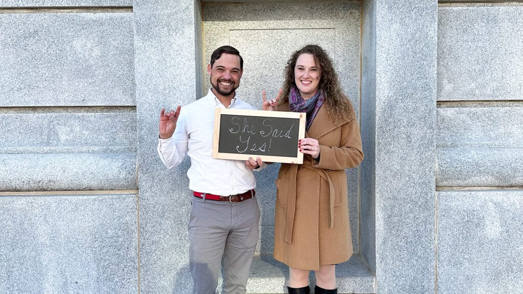 A newly engaged couple poses in front of the Memorial Belltower with a sign that says "She said yes."