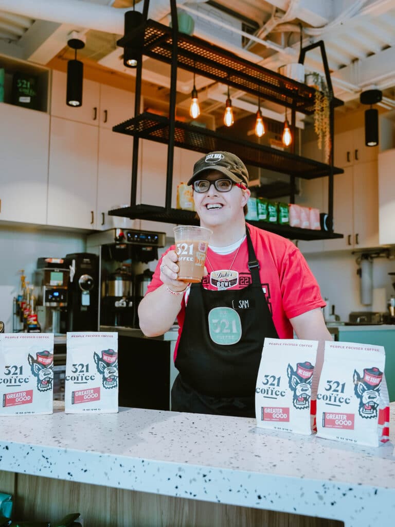 321 employee displays a fresh cup of coffee behind a row of NC State x 321 branded coffee.