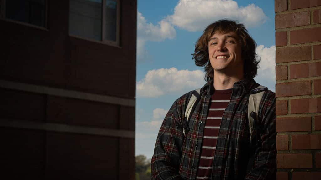 Portrait of Galen Wilcox smiling as he stands beside a brick wall against a blue-sky background.