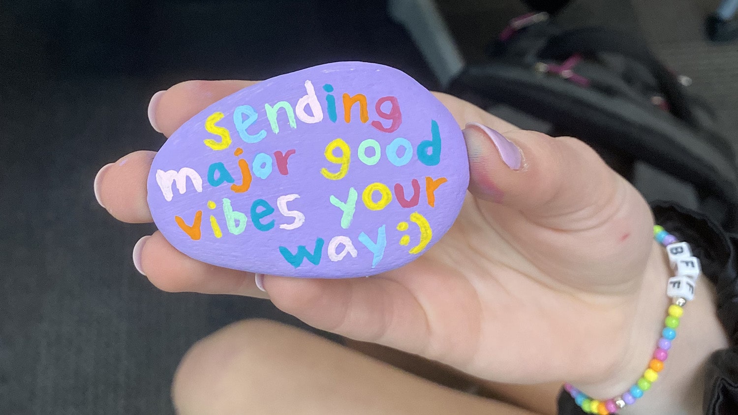 A student holds a painted purple rock with the message "sending major good vibes your way :)" on it. Each letter is in a different color. This photo is a close-up of the rock and the student's hand.