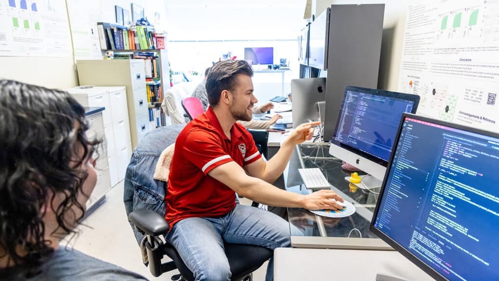 Jacob Gerlach sits at a computer and points to a line of data on the screen. Another person sits to his right, looking at his computer screen.