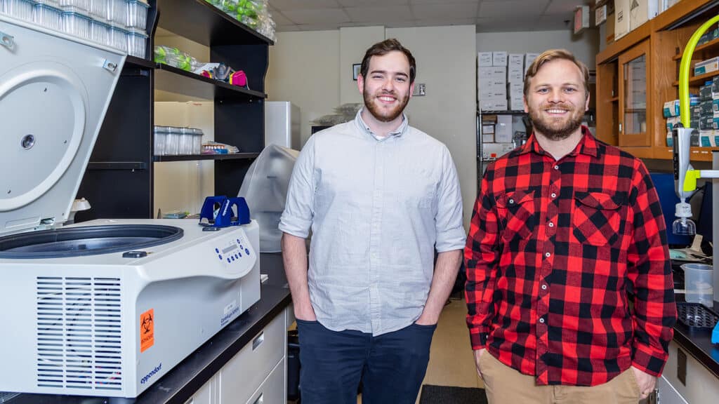 Thane Jones and Josh Harris stand next to each other for a portrait inside  Collaborations Pharma’s lab. They are standing next to lab equipment.