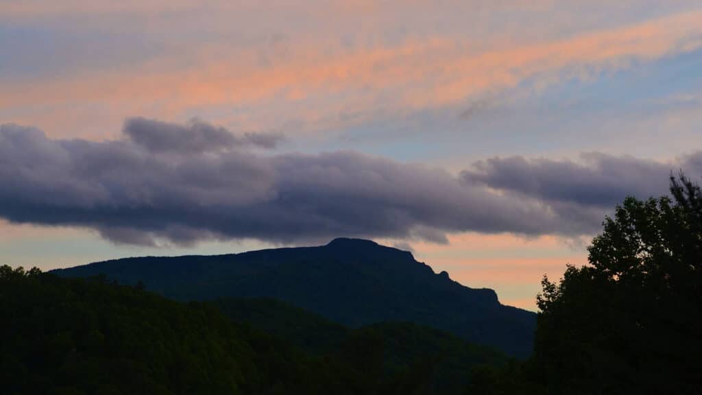 Grandfather Mountain looms in the distance at dusk, with anthropomorphic features that appear to stare stoically up at a pink and blue, cloud-wisped sky.