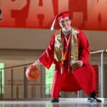 Billy Fryer dribbles a basketball inside Reynolds Coliseum while wearing his red cap and gown.