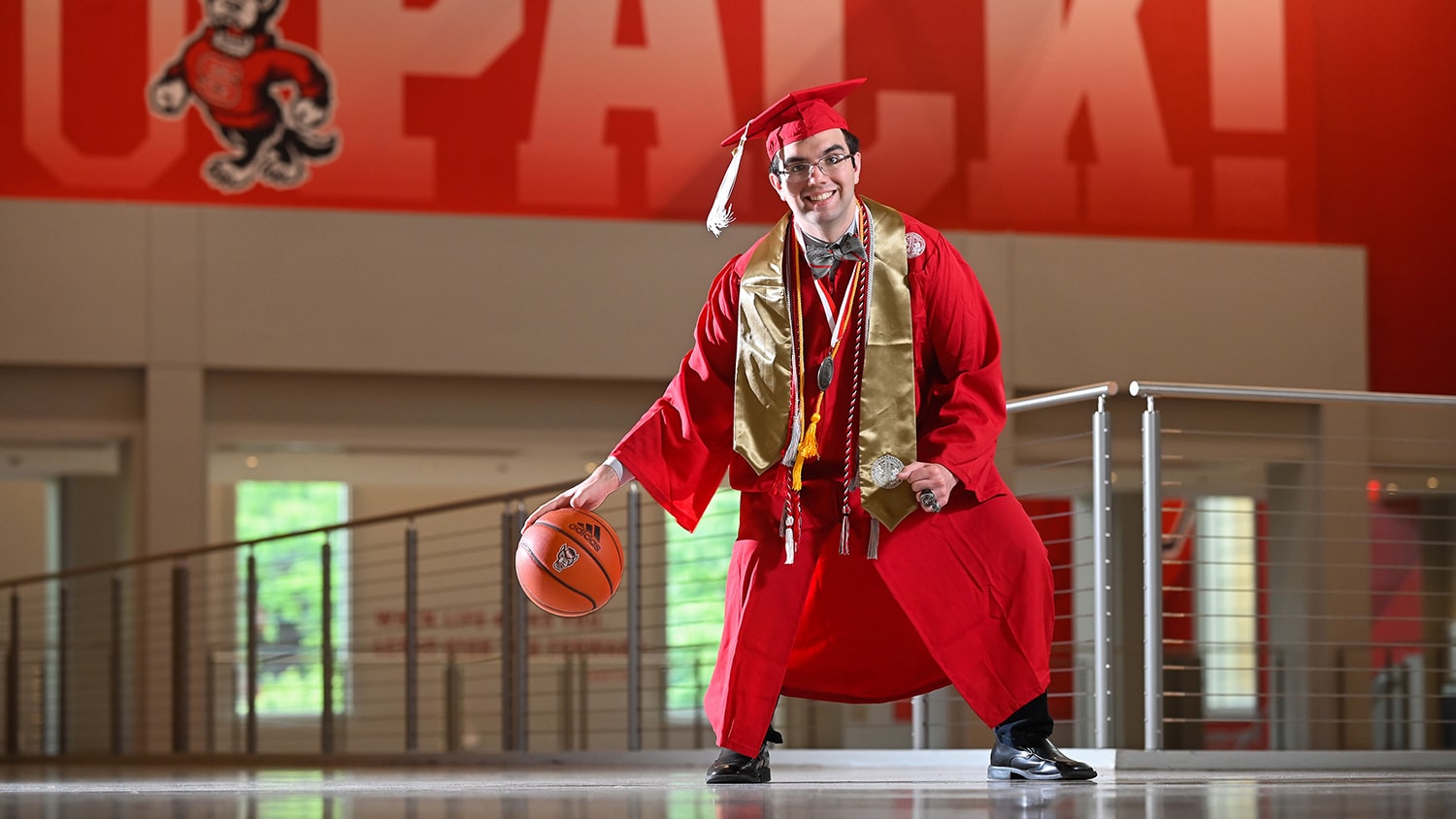 Billy Fryer dribbles a basketball inside Reynolds Coliseum while wearing his red cap and gown.