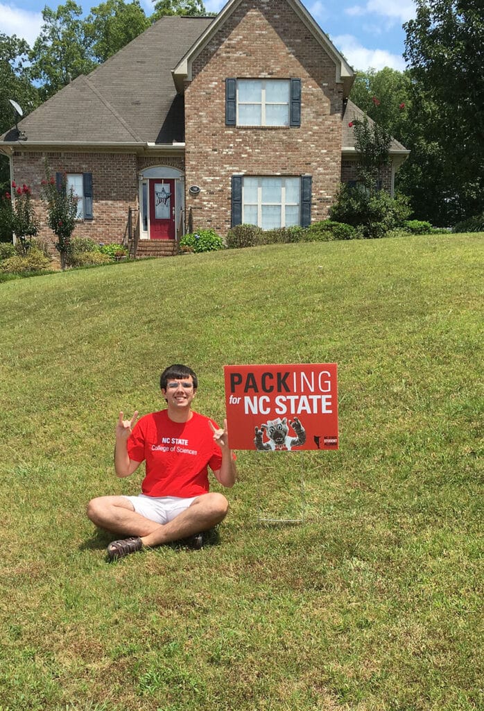 Billy Fryer sits in front of his house with a yard sign that reads "Packing for NC State"