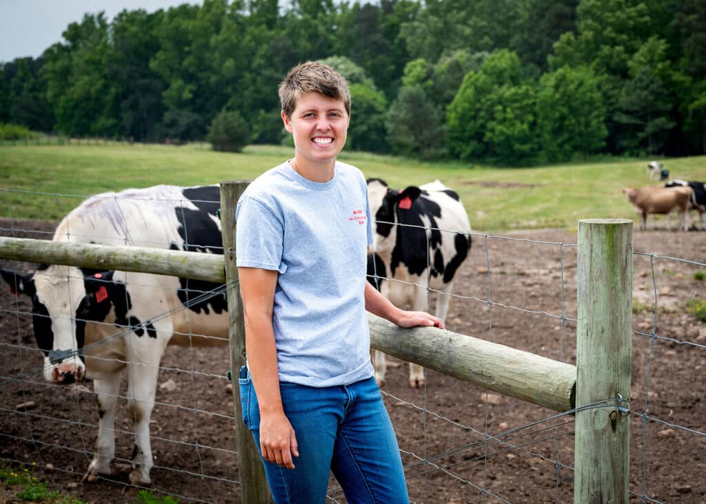 Caroline Vande Berg stands in front of a fence at NC State's Dairy Research and Teaching Farm. Heifers stand in the background, roaming in a field.