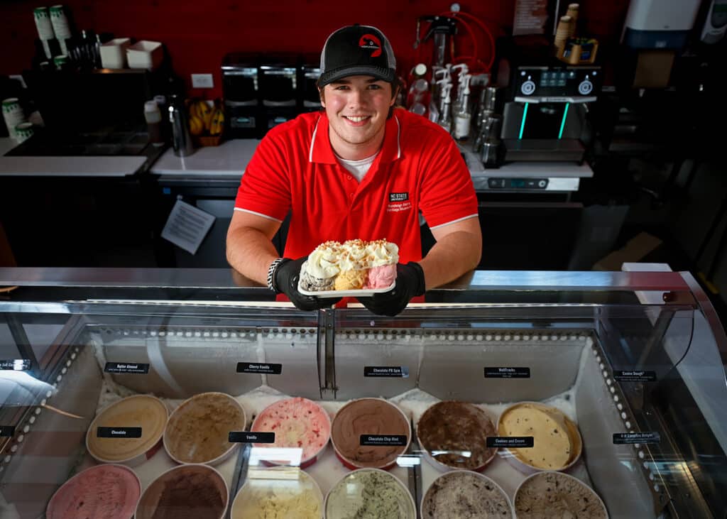 Jordan Stanley stands behind the ice cream counter at the Dairy Education Center and Creamery. He holds a sundae with three flavors of ice cream and whipped cream.
