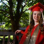 Kelsey O'Connor sits outside on a swinging bench wearing her red cap and gown.