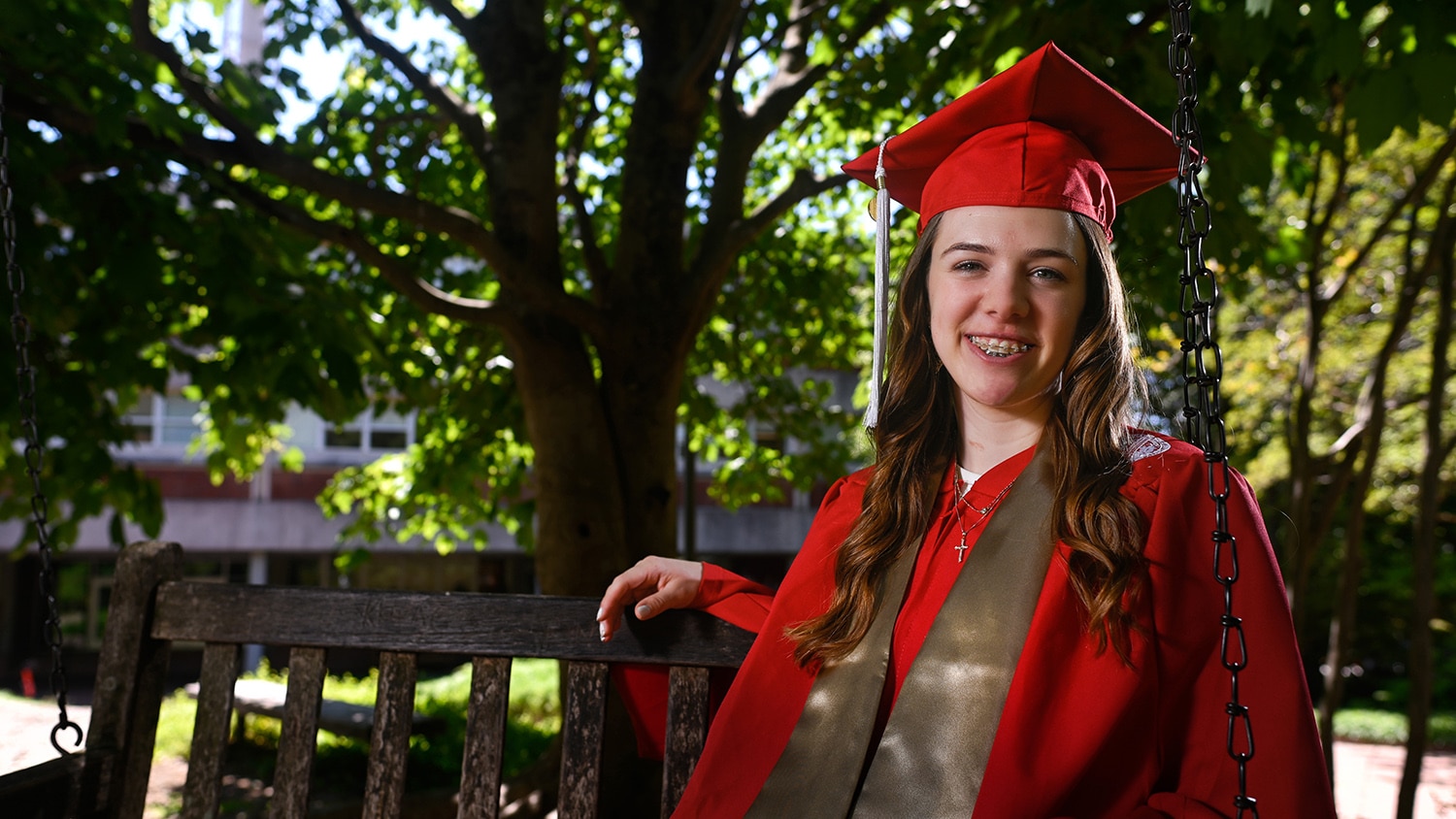 Kelsey O'Connor sits outside on a swinging bench wearing her red cap and gown.