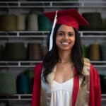 Ritika Shamdasani stands in front of shelves holding spools of yarn wearing her red cap and gown.