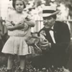 A black-and-white photo shows a young Mary Yarbrough, appearing to be of early grade-school age, standing alongside her kneeling father in a yard filled with flowers in early bloom. They each hold a duck in their hands.