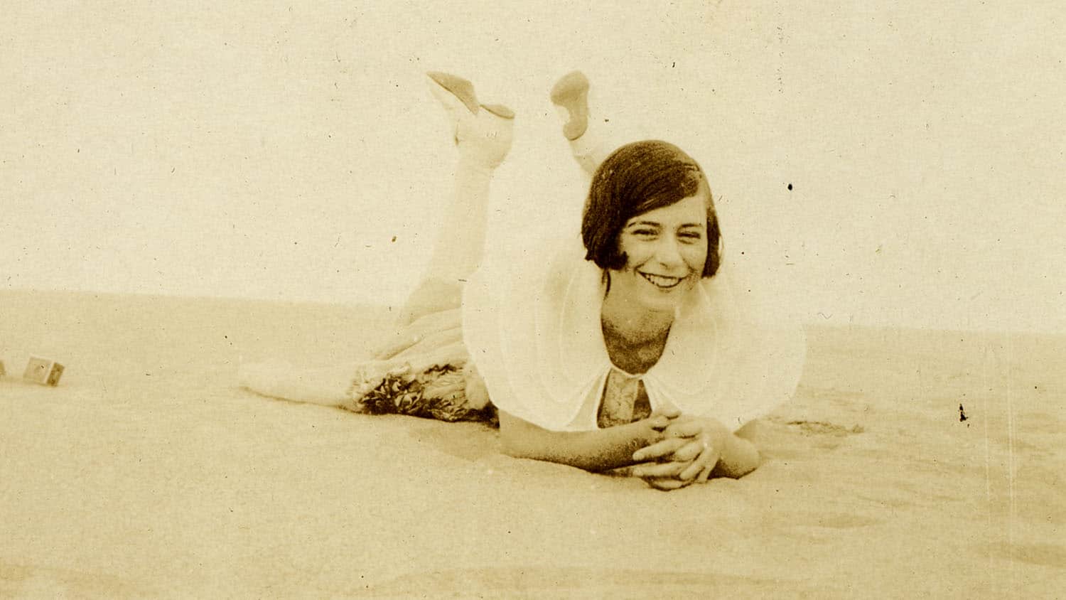 A sepia-toned photos shows a late adolescent Mary Yarbrough smiling at the camera as she lays on her stomach atop a sand dune at Kill Devils Hills in North Carolina's Outer Banks.