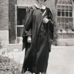 A black-and-white photos shows a young Mary Yarbrough standing in dark-colored graduation attire in front of a brick building on the campus of Meredith College. She cradles her diploma in one arm.
