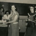A black-and-white photos shows an adult Mary Yarbrough standing while looking back at the camera in a laboratory classroom. She is surrounded by smiling students as a subtle smile plays across her face.