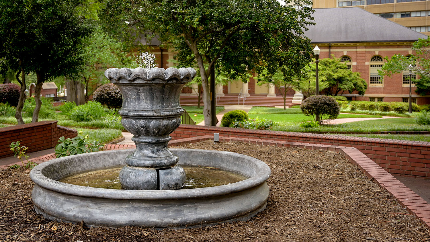 A fountain erupts with water in the foreground at Mary Yarbrough Court, with trees and greenery in the background.