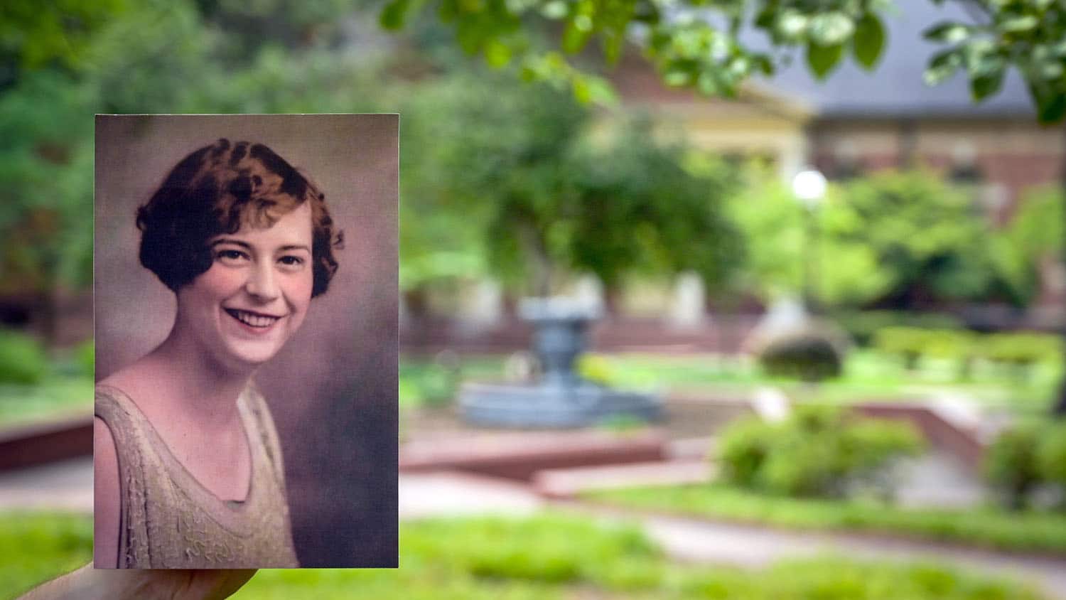 A colorized photo of a young adult Mary Yarbrough is held up in front of a swath of greenery, brick pathways and a fountain at Yarbrough Court.