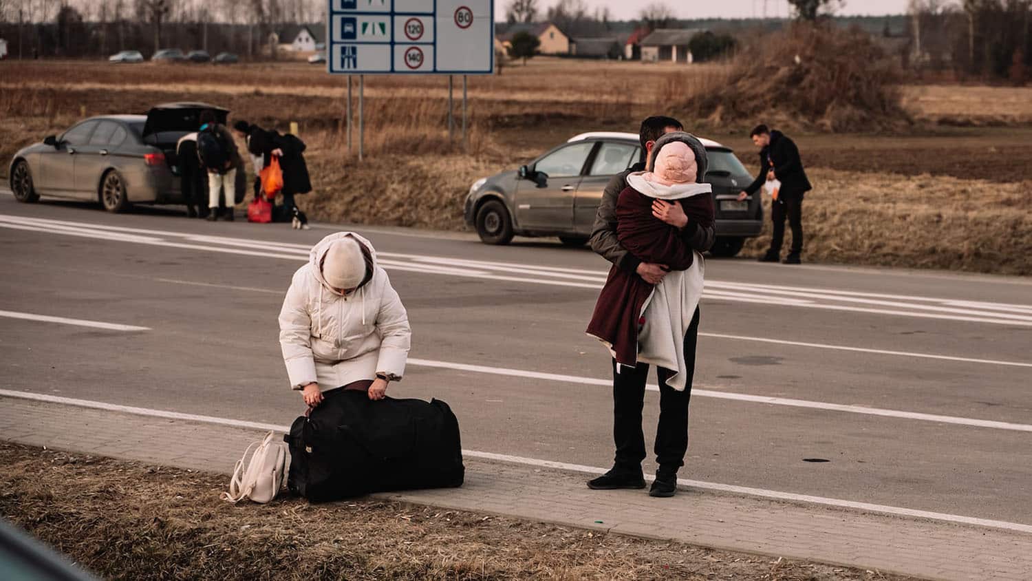 two adult Ukrainian refugees stand by a roadside. One of them is holding a child. The road signs tell you that the scene is inside the European Union.