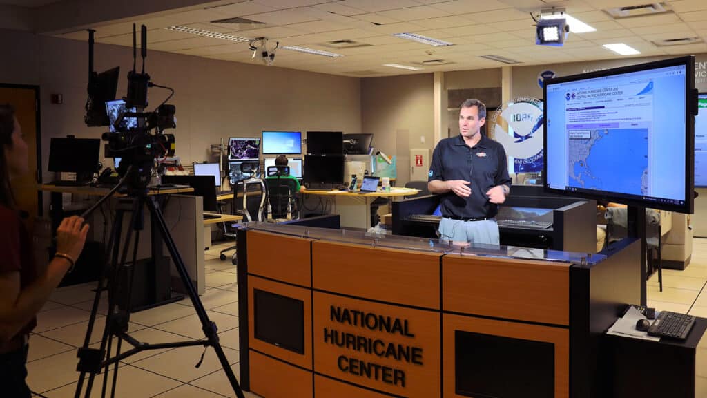 Michael Brennan stands in front of a screen inside a broadcast studio. A TV camera and camera operator are recording him.