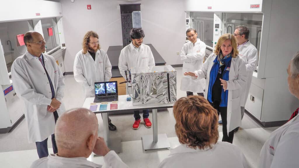 A group of researchers in white coats — engineers and scientists — convene to discuss a project in the Organic and Carbon Electronics Laboratories.