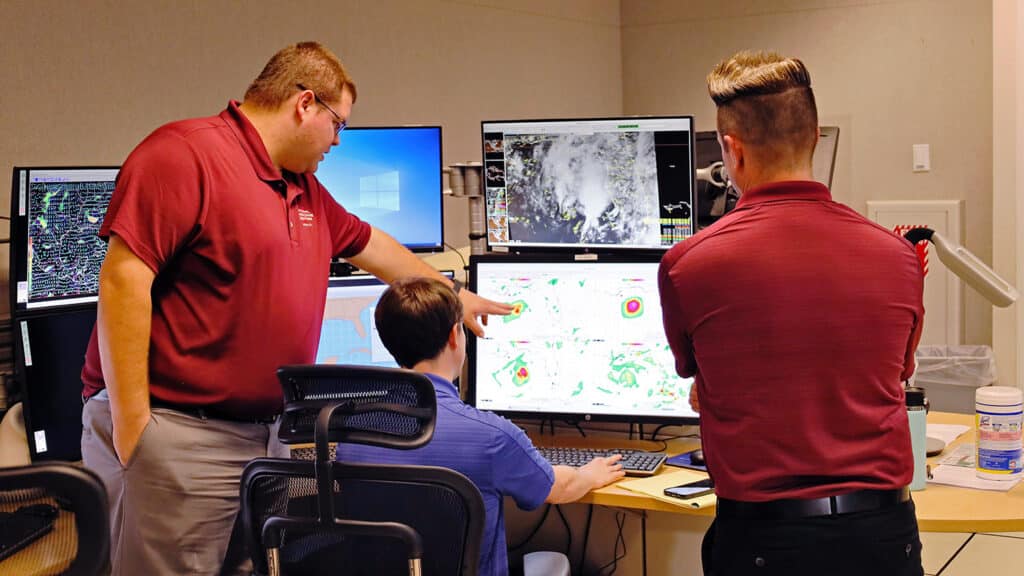 Three people gather around five computer monitors, each showing different maps and data of an approaching storm.