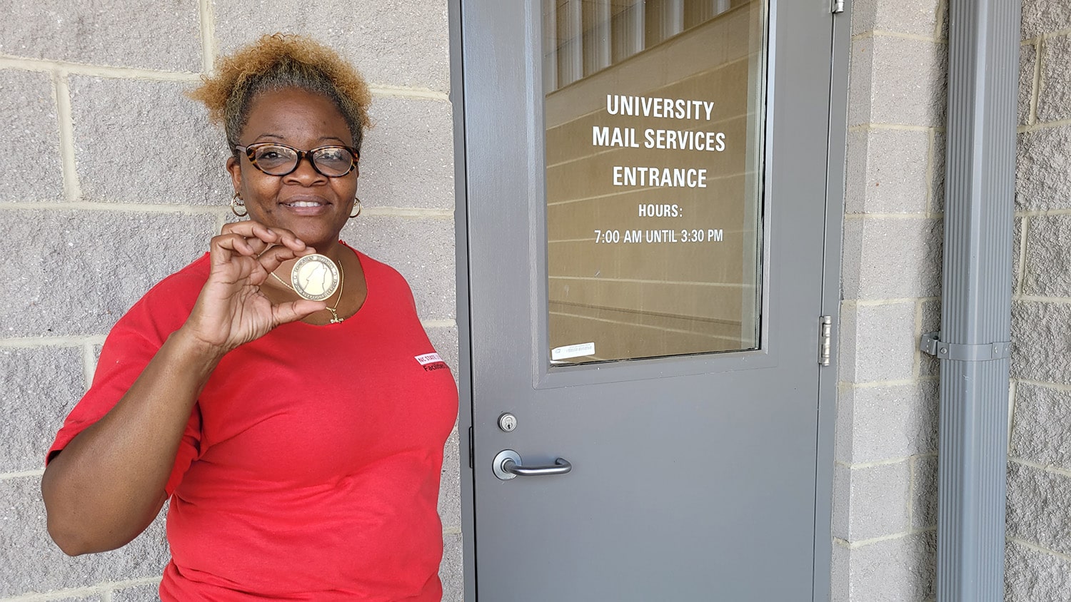 Ebony Leggett holds up her Facilities Coin for Exceptional Service next to the door for the Mail Services office.