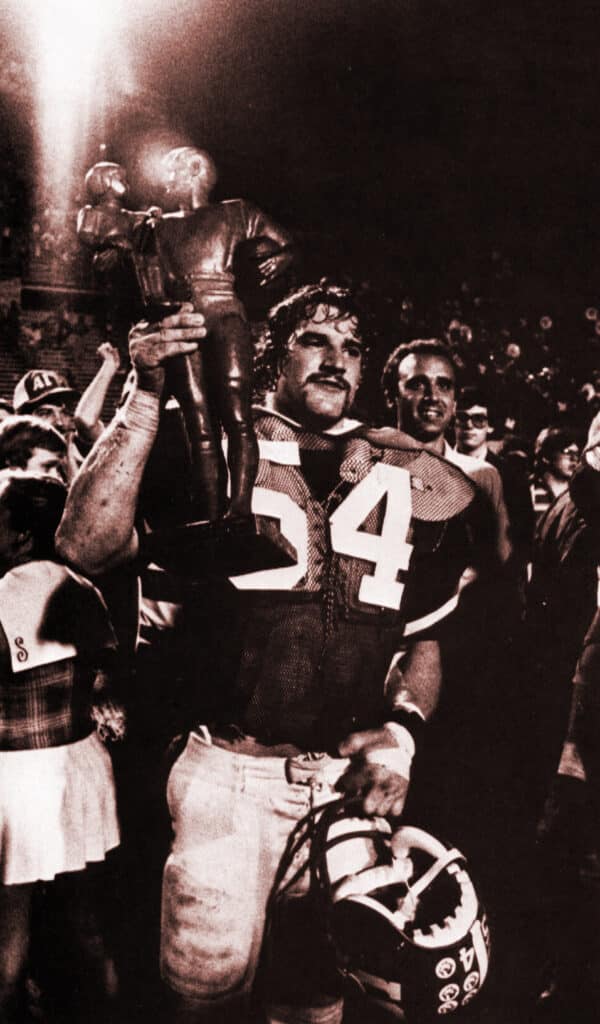 Bill Cowher in a black and white photo, dressed in an NC State jersey and holding a trophy in a crowd of people