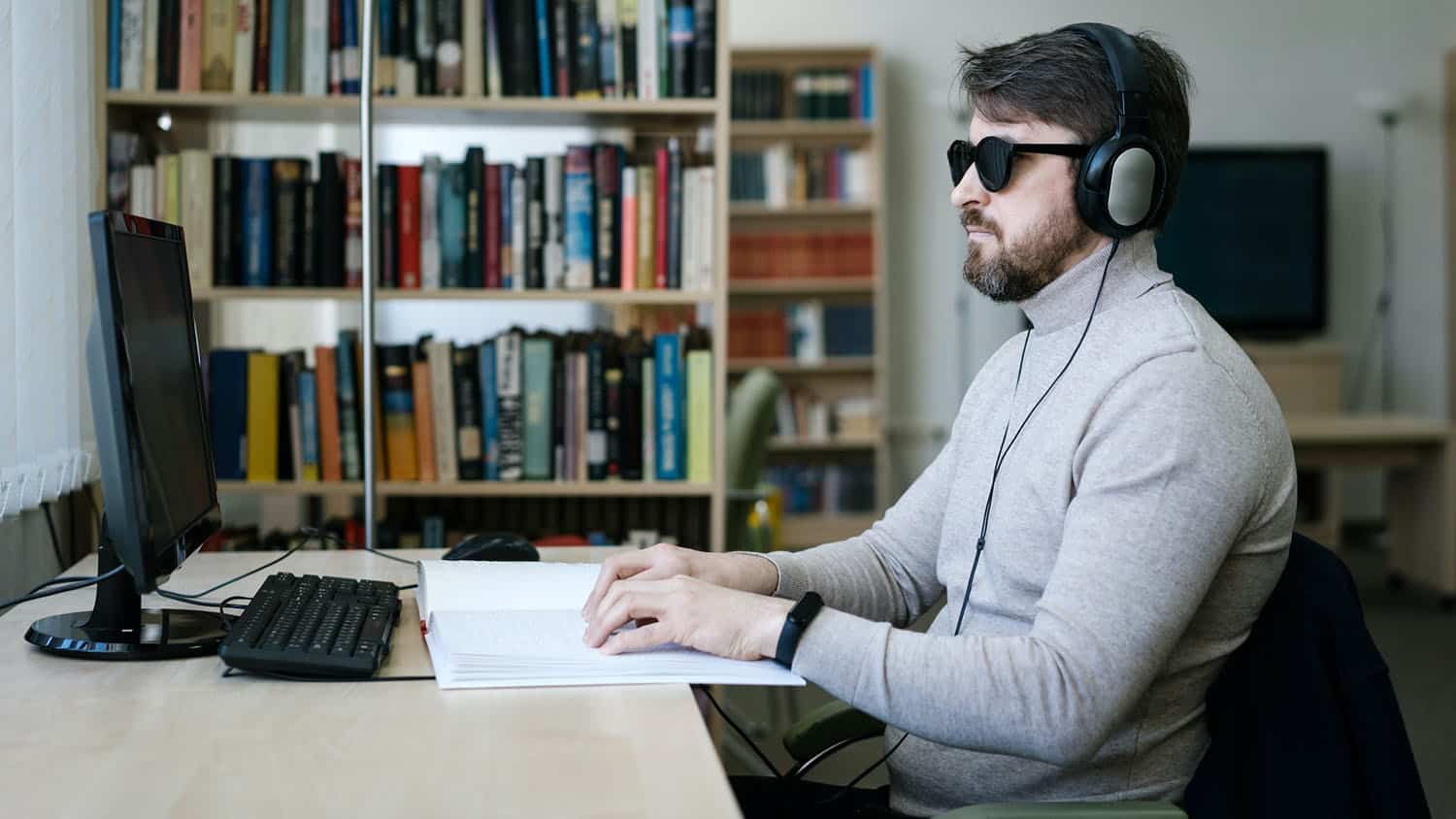 a blind man sits at a desk with a computer. he is wearing headphones and reading a book written in braille.