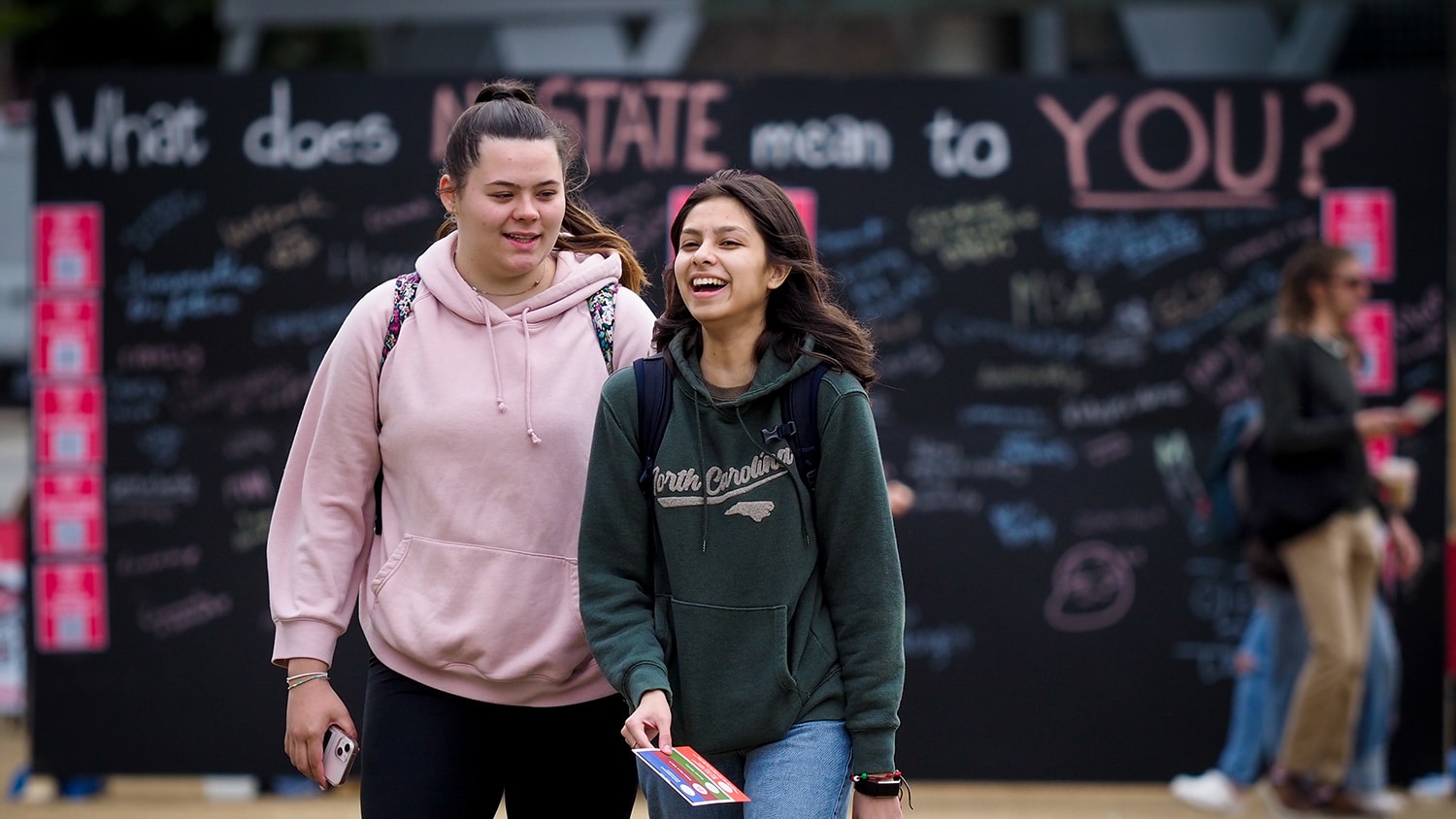 Two students laugh together while walking in front of a chalkboard sign that reads "What does NC State mean to you?" with hand written comments below the question.