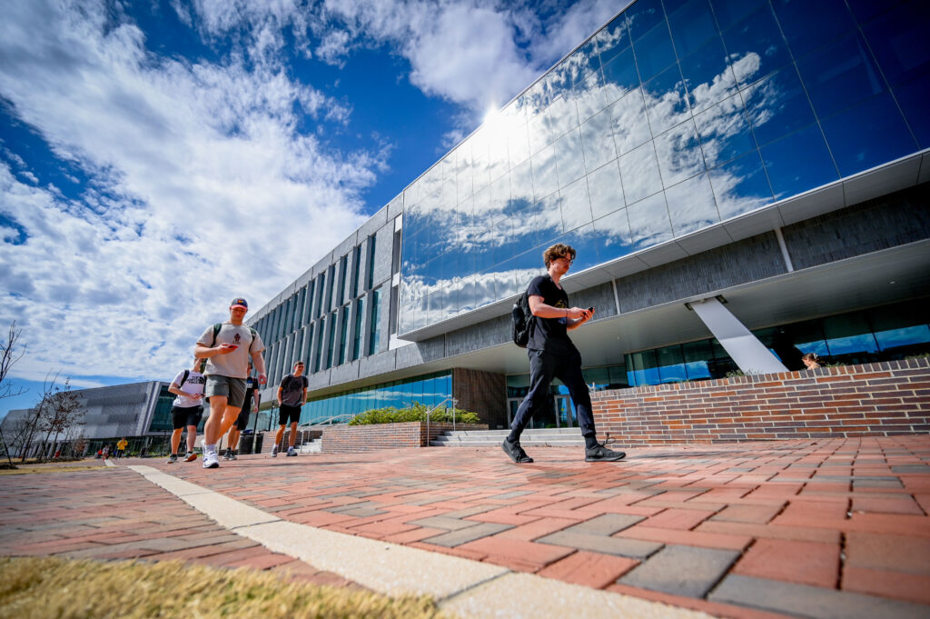 Students walk past Fitts-Woolard Hall on Centennial Campus. Clouds reflect off the windows of the building facade.