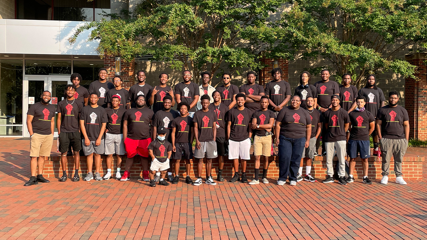 A large group of male students in black T-shirts stand on a brick path at NC State