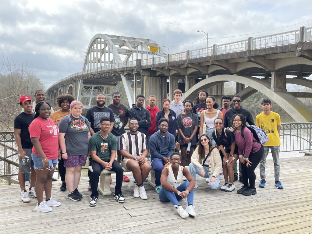 A large group of NC State students on a wooden board walk, with the Edmund Pettus Bridge in the background. 