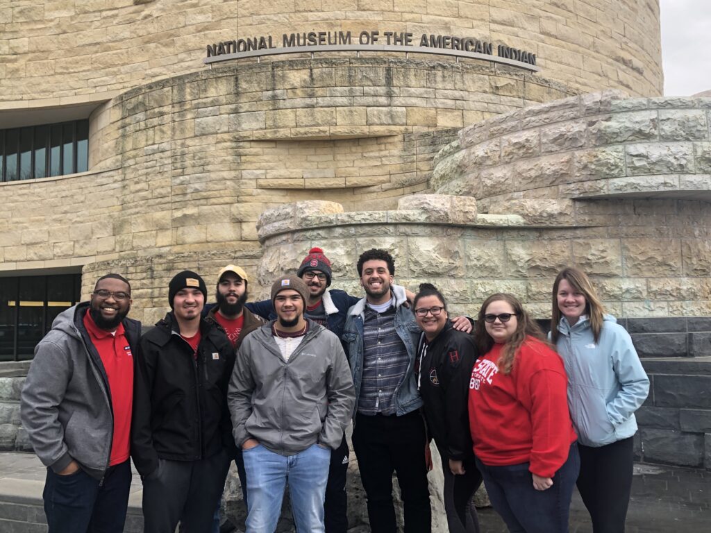 A group of people in red and black NC State clothing stand in front of a building with a sign that reads "National Museum of the American Indian."