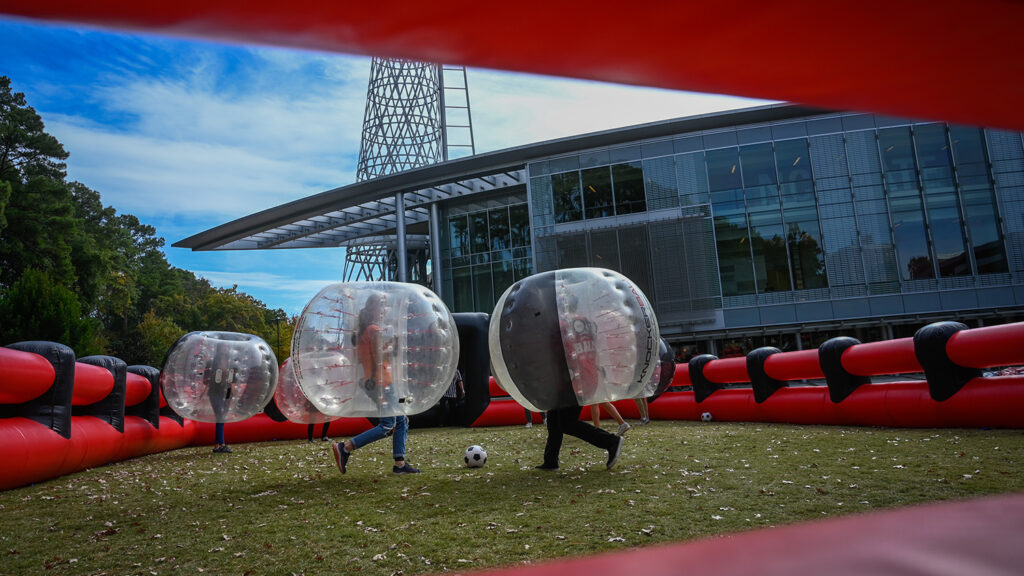 Students chase a soccer ball outside while wearing large inflatable balls. 