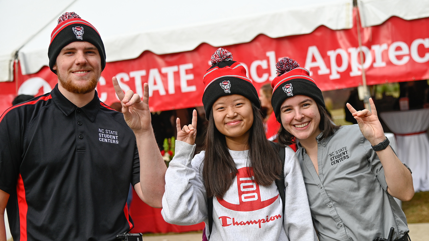 Two employees and a student, wearing matching NC State beanies, pose outside with wolfies up during Pack Appreciation Day.