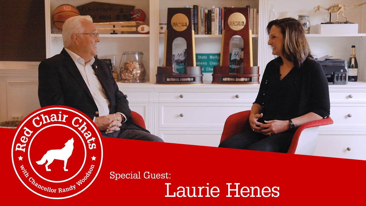 Chancellor Woodson interviewing Coach Laurie Henes sitting in her home in front of two NCAA trophies. The image also contains the Red Chair Chats graphic, which says "Red Chair Chats with Chancellor Woodson. Special guest: Laurie Henes"